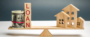 miniature wooden houses balance on a scale with blocks spelling LOAN and a bundle of money on the other side